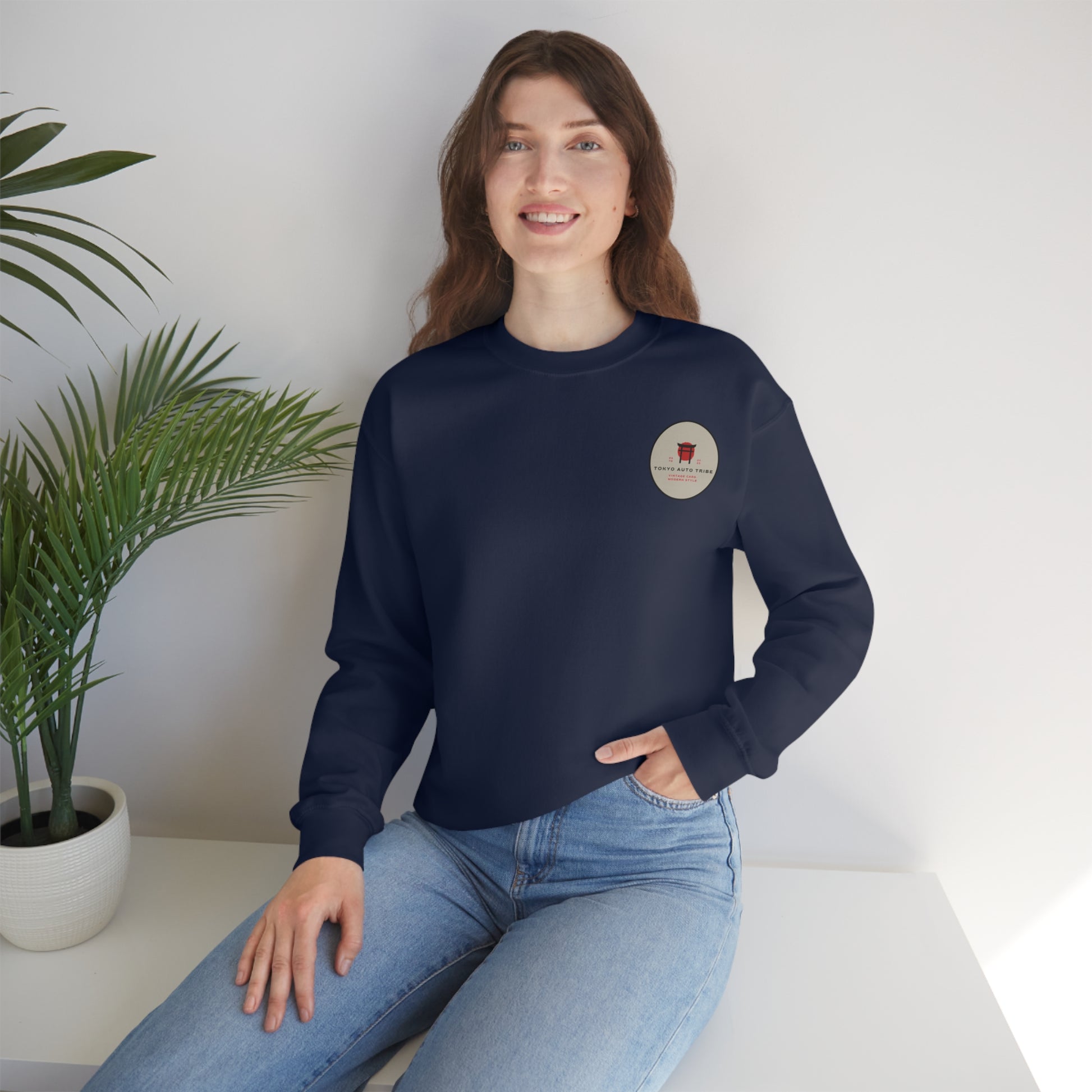 Smiling woman wearing a navy blue Wagon Chibi Sweater with a small graphic emblem on the chest, seated indoors near a green potted plant in casual blue jeans.