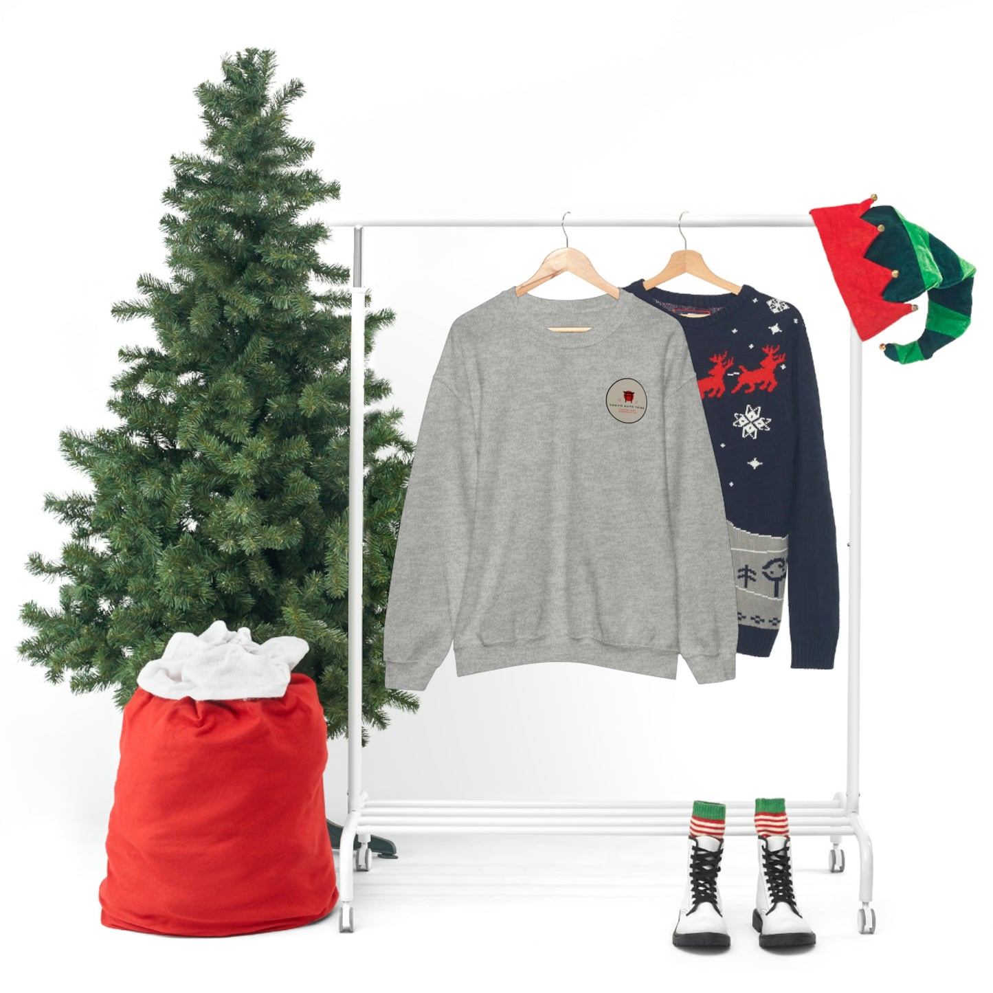 Two festive sweaters hanging on a white clothing rack beside a Christmas tree and red sack, featuring The Family Wagon Sweater in gray alongside a holiday-themed navy sweater with reindeer.