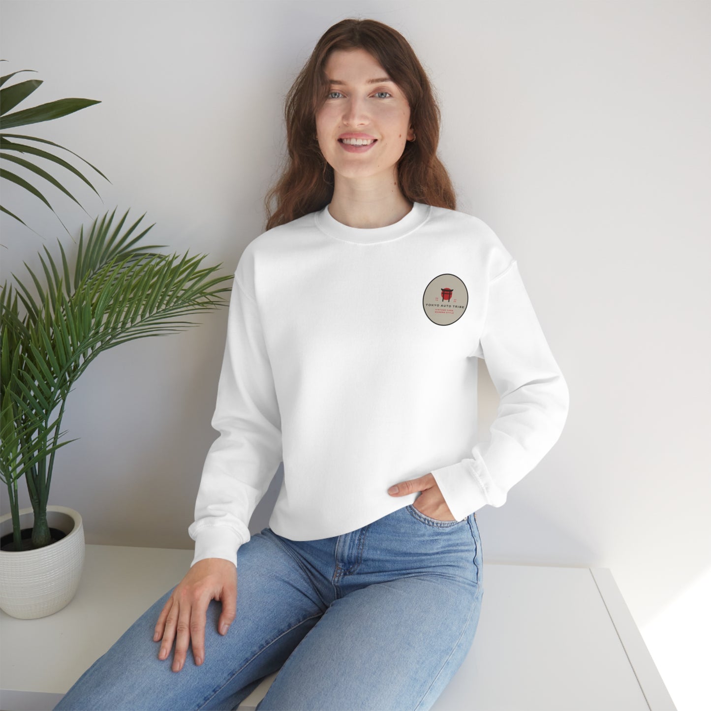 Smiling woman wearing a white Wagon Chibi Sweater with a small vintage-inspired graphic on the chest, sitting casually by a white surface next to a green potted plant.
