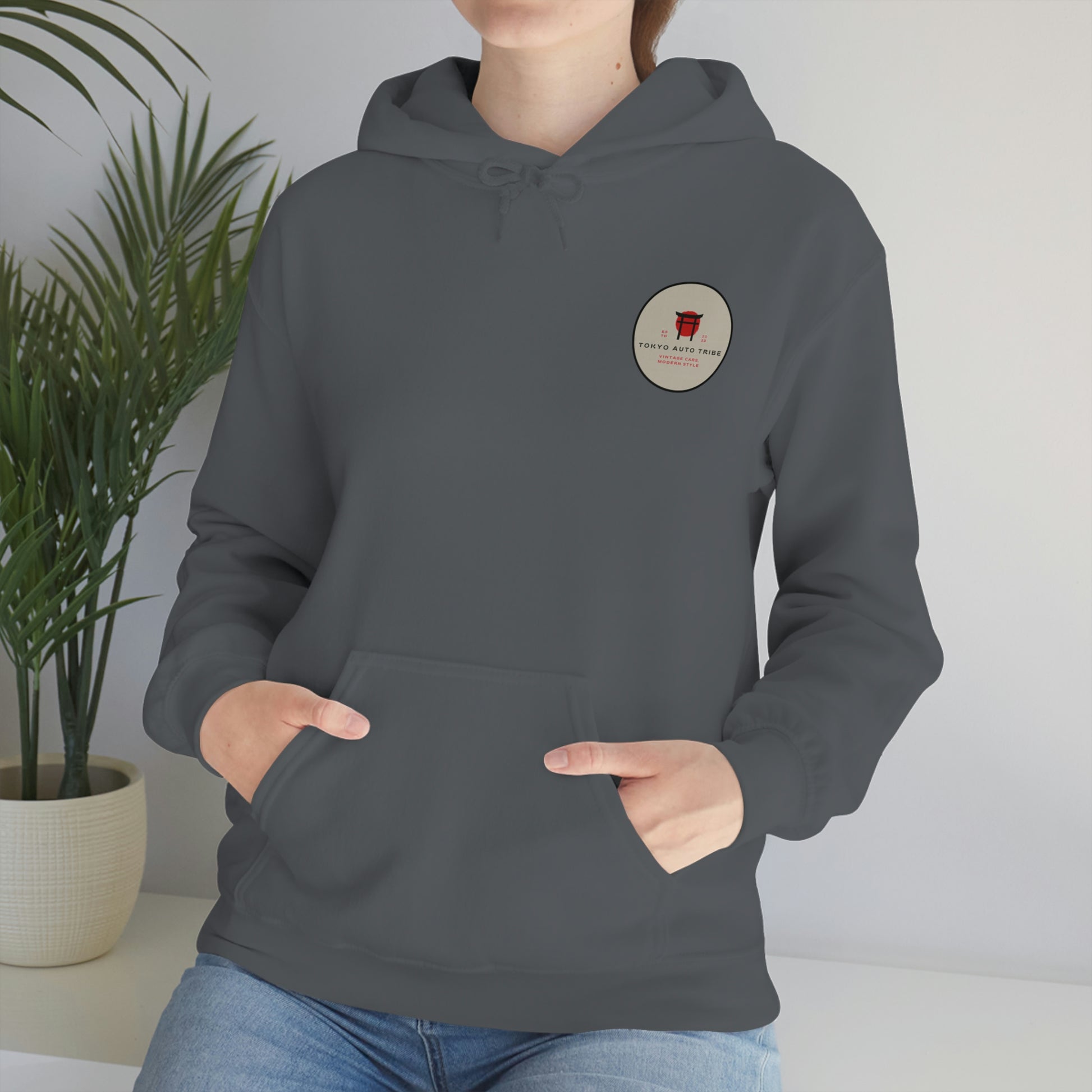 Person wearing a dark gray Groovy Grocery Runner Hoodie with a small round graphic on the chest, posing indoors near a green leafy plant, showcasing casual automotive streetwear inspired by Japanese car culture.