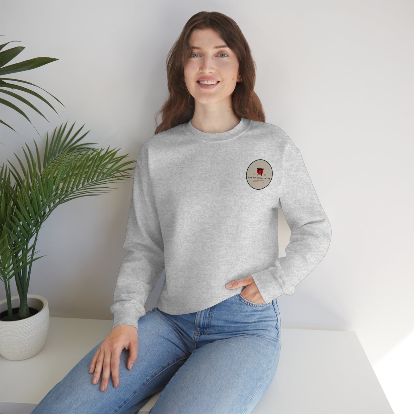 Young woman wearing a light gray Wagon Chibi Sweater with a small graphic on the chest, paired with blue jeans, sitting indoors next to a potted plant.