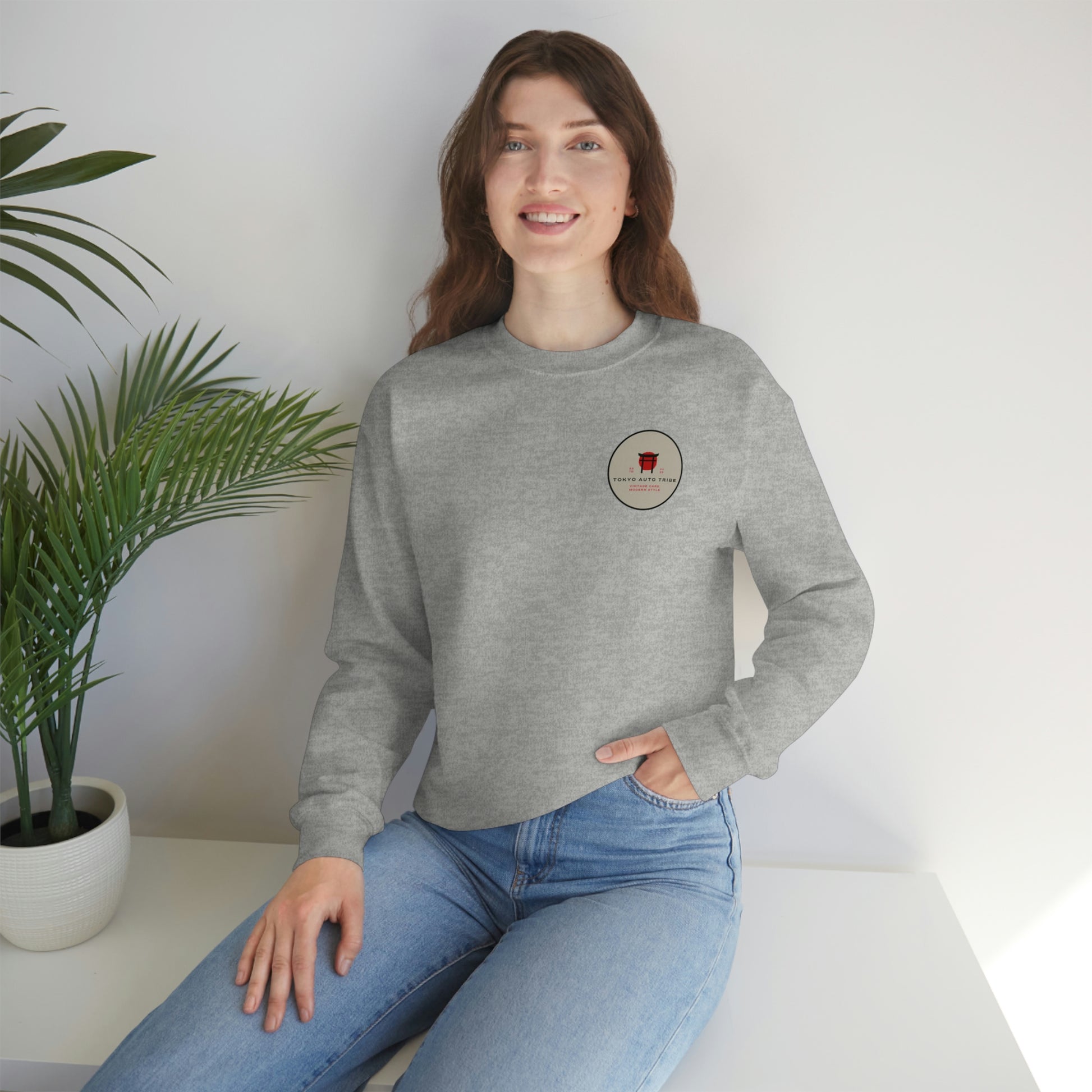 Smiling woman wearing a light gray crewneck sweatshirt with a small round logo on the chest, styled casually with blue jeans and posed indoors by a green potted plant in The Family Wagon Sweater.