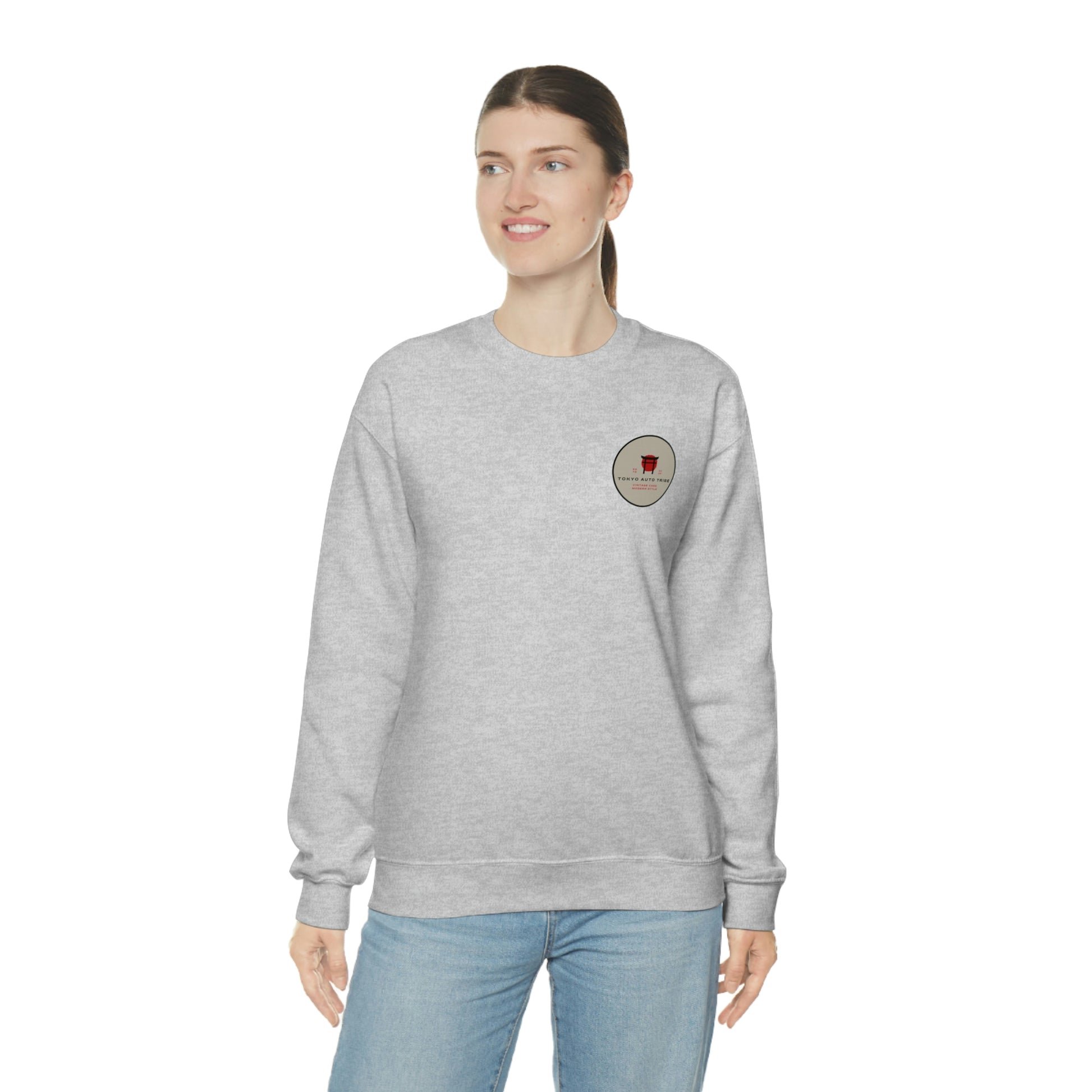 A woman wearing a light gray crewneck sweatshirt featuring a small circular graphic on the chest, styled as The Family Wagon Sweater for fans of Japanese car culture and late-night drives.