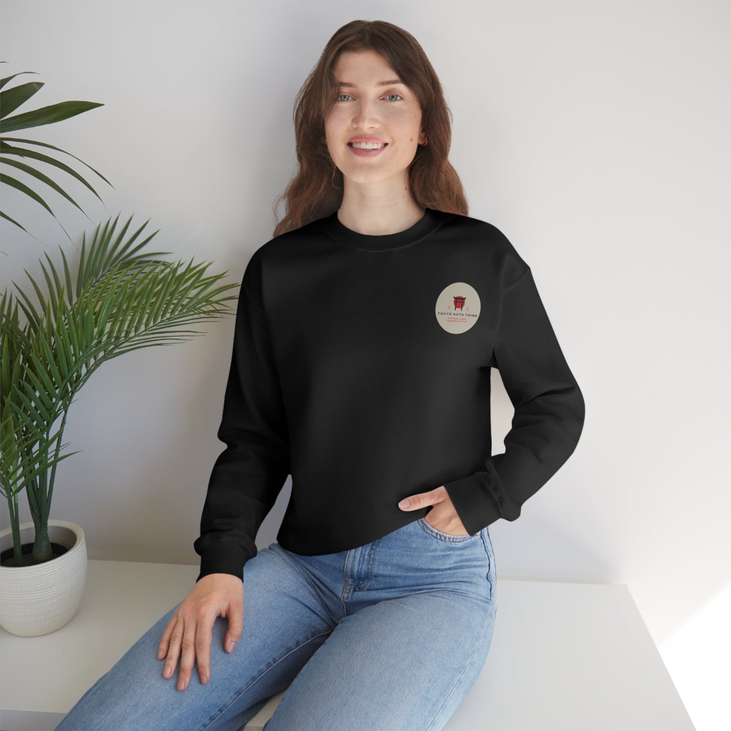 Woman wearing a black Wagon Chibi Sweater featuring a small red graphic and text on the chest sitting casually beside a potted plant in a bright, minimalist room.