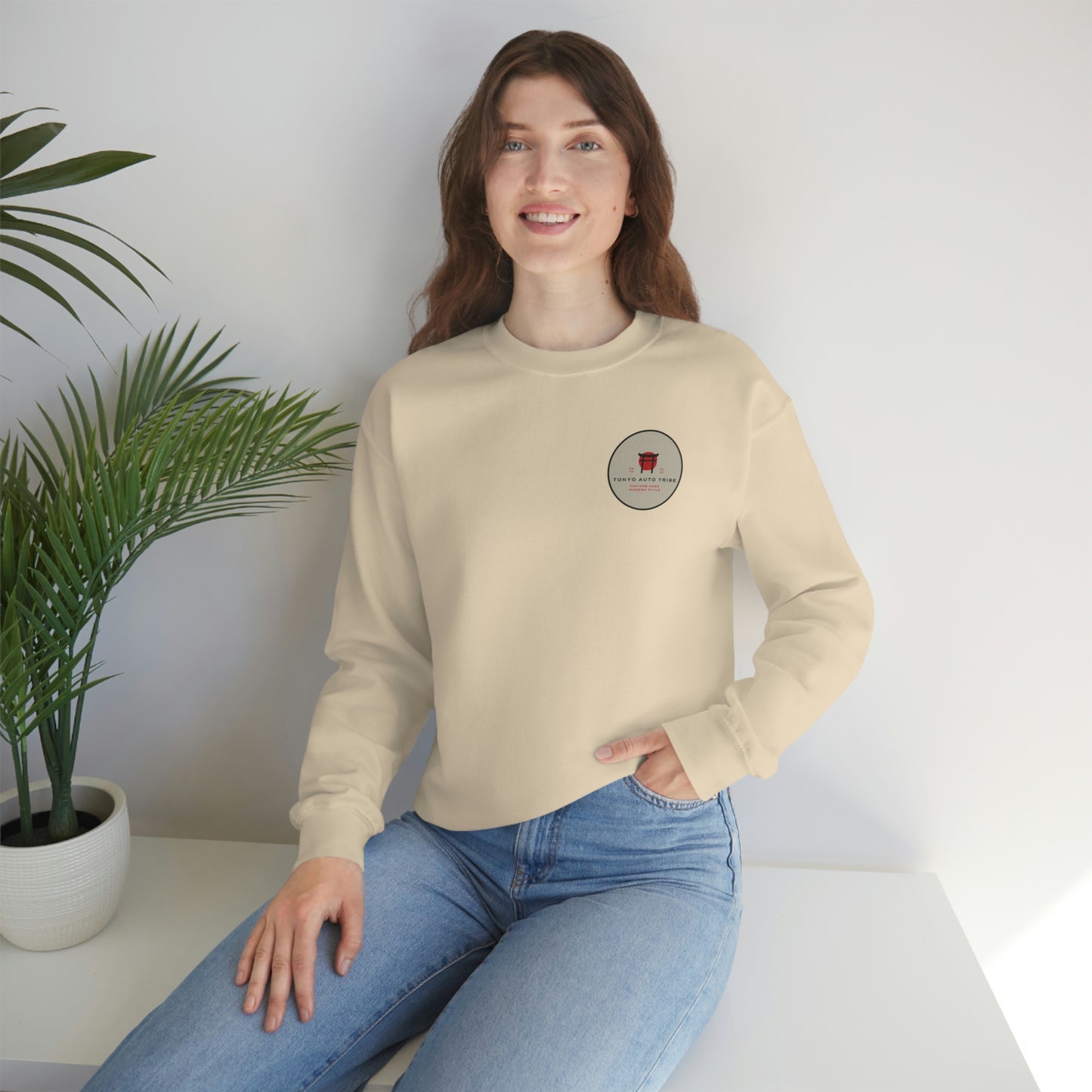 A woman wearing The Family Wagon Sweater in beige, featuring a small round graphic on the chest, styled casually with light blue jeans in a bright, minimalist setting.