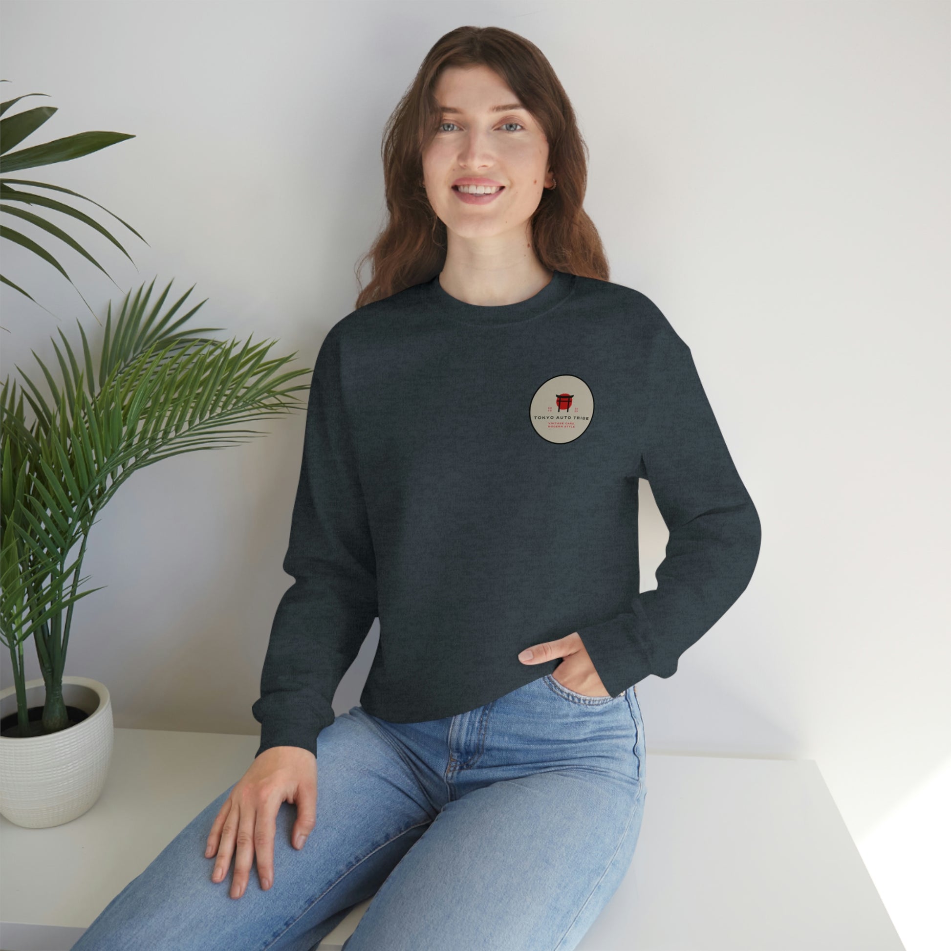 Woman sitting indoors wearing a dark sweatshirt featuring a small circular logo on the chest, styled casually with light blue jeans, showcasing The Family Wagon Sweater from Tokyo Auto Tribe inspired by late-night drives and Japanese car culture.