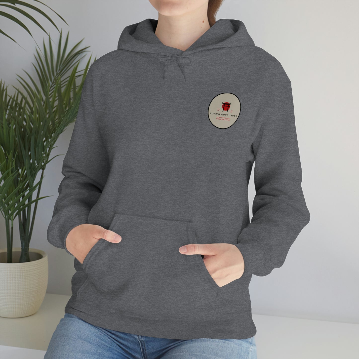 Person wearing a dark gray Groovy Grocery Runner Hoodie with a small circular logo on the chest, showcasing casual Japanese car culture-inspired streetwear in a relaxed indoor setting.