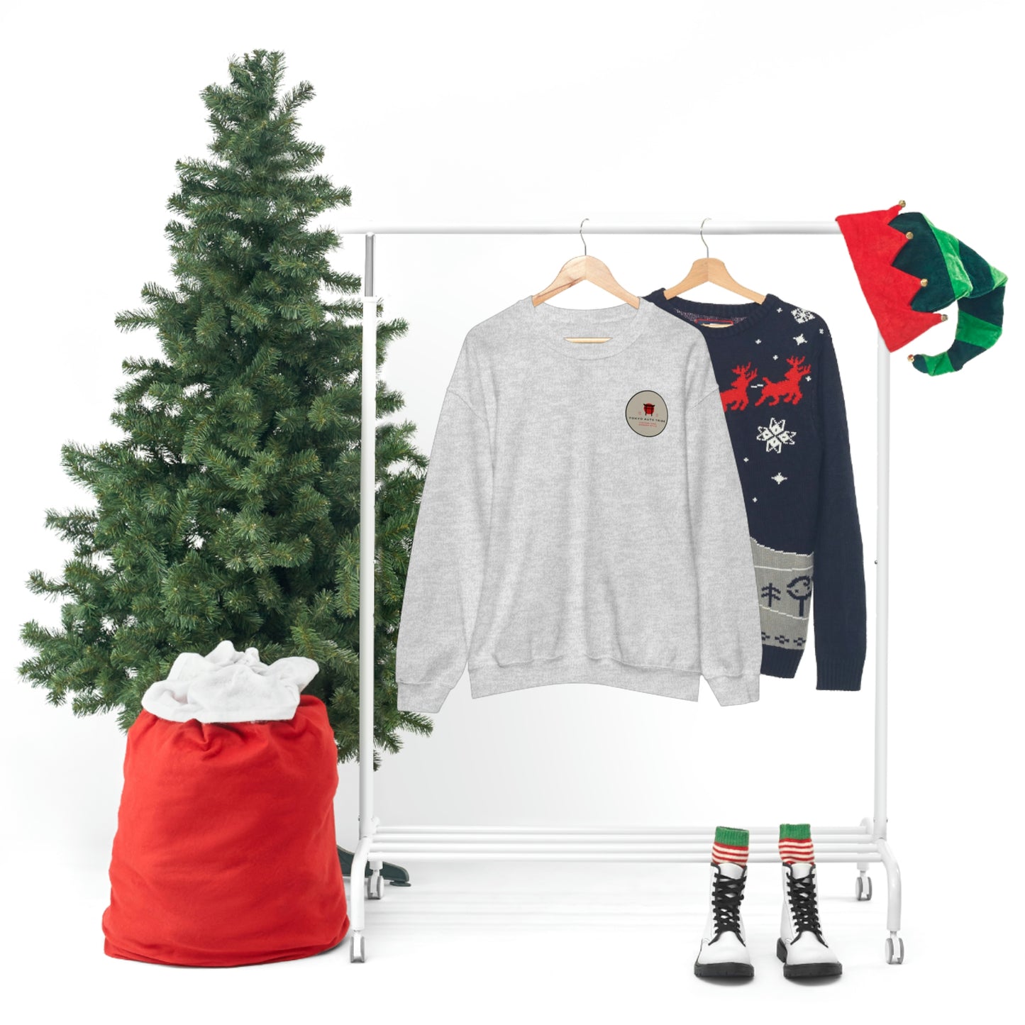 The Family Wagon Sweater displayed on a hanger beside a festive Christmas sweater, with a green Christmas tree, red sack, elf hats, and striped socks with black boots creating a holiday-themed setting.