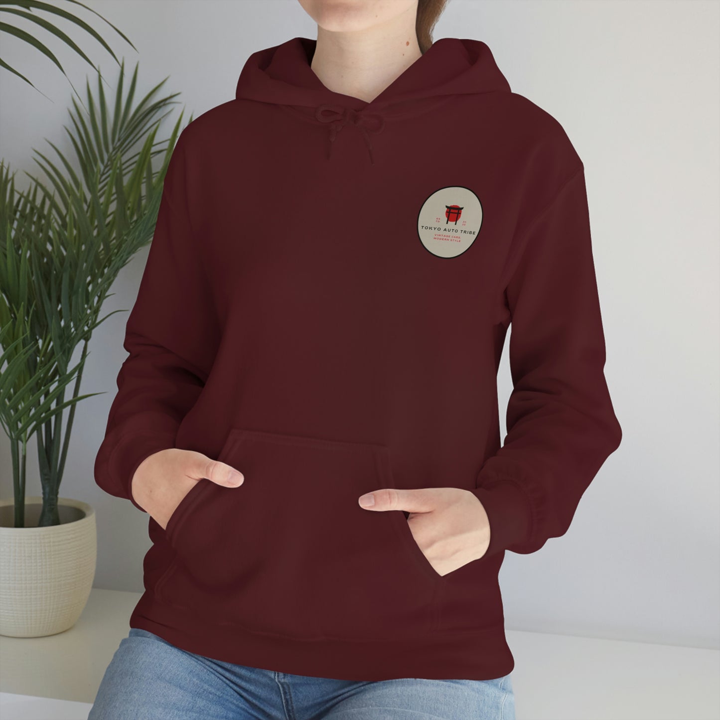 Person wearing a maroon Groovy Grocery Runner Hoodie with a small round graphic logo on the chest, styled casually with hands in front pocket, set against a minimalist background with a green plant.