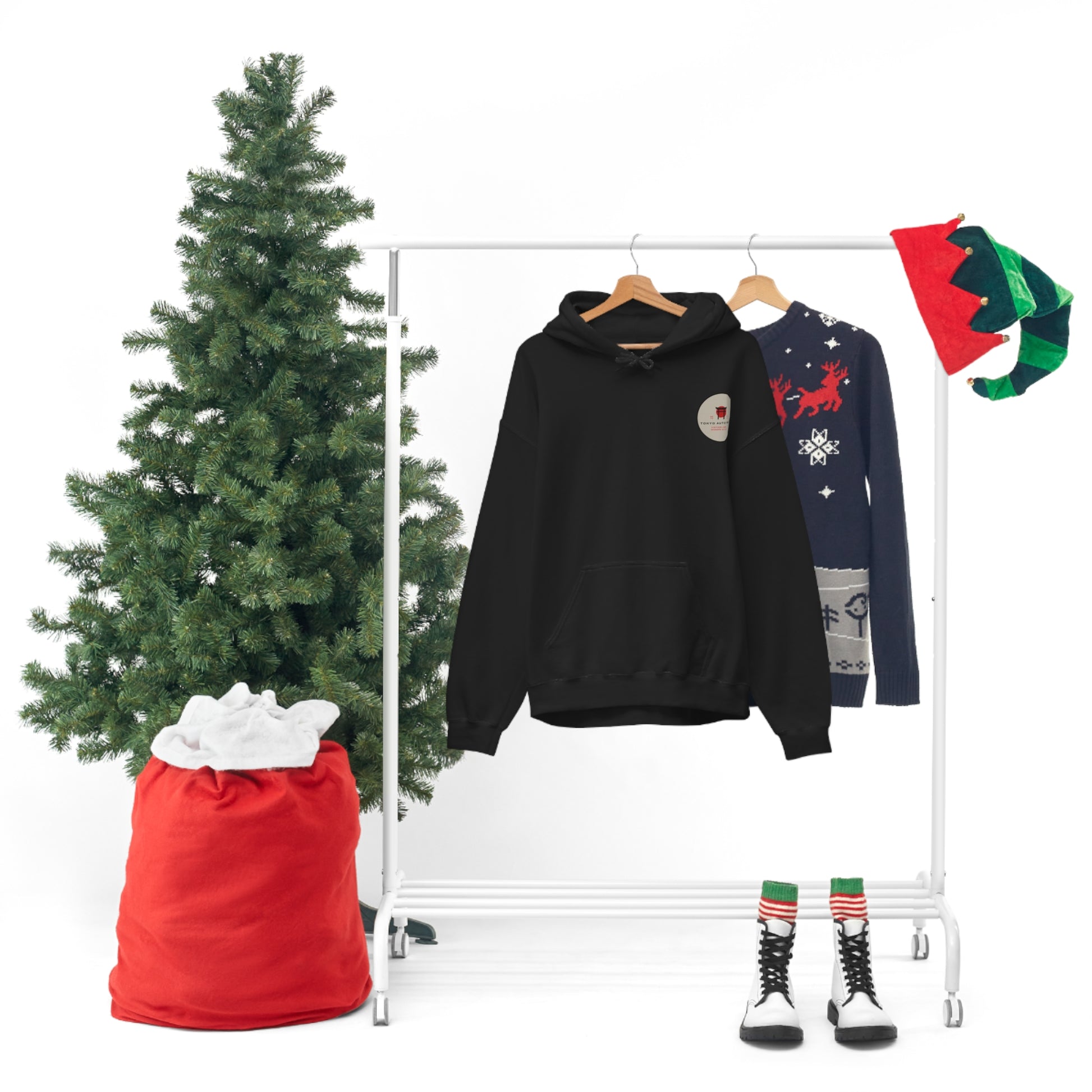 Black Wagon Chibi Hoodie hanging on a rack beside a festive Christmas sweater, seasonal elf hats, boots with striped socks, a decorated Christmas tree, and a red sack, capturing a cozy holiday vibe.