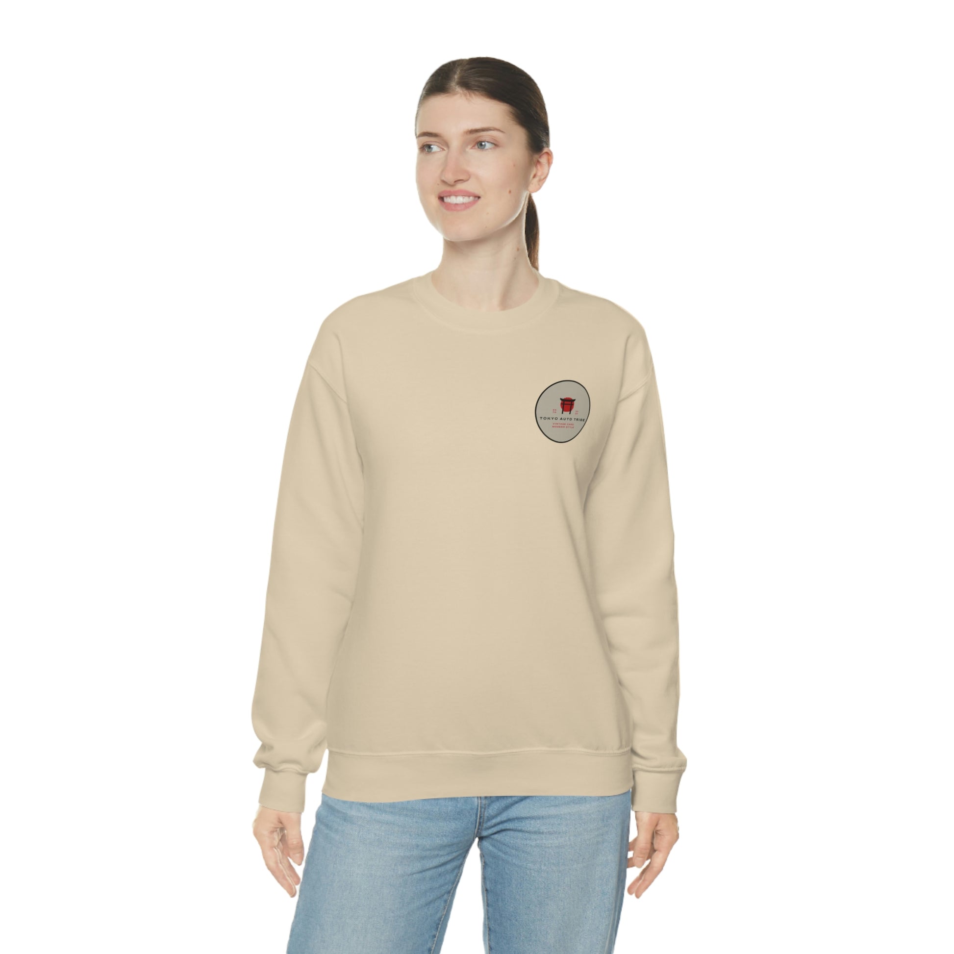 Woman wearing a beige sweatshirt featuring a small graphic emblem on the chest, styled casually with jeans, showcasing The Family Wagon Sweater inspired by Japanese car culture.
