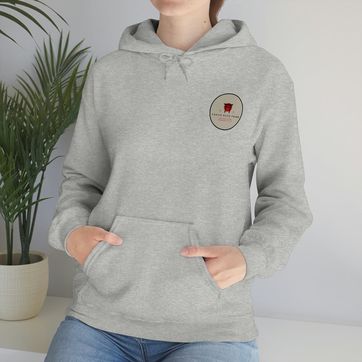 Person wearing a light gray Groovy Grocery Runner Hoodie with a small circular logo on the chest, standing beside a potted plant in a bright indoor setting.