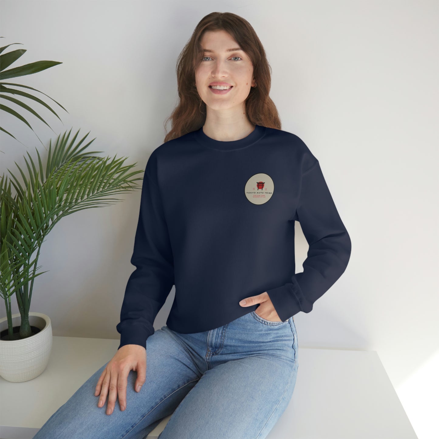 A smiling woman wearing The Family Wagon Sweater in navy blue, styled casually with light blue jeans, sitting indoors near a green potted plant, embodying relaxed automotive streetwear inspired by Japanese car culture.