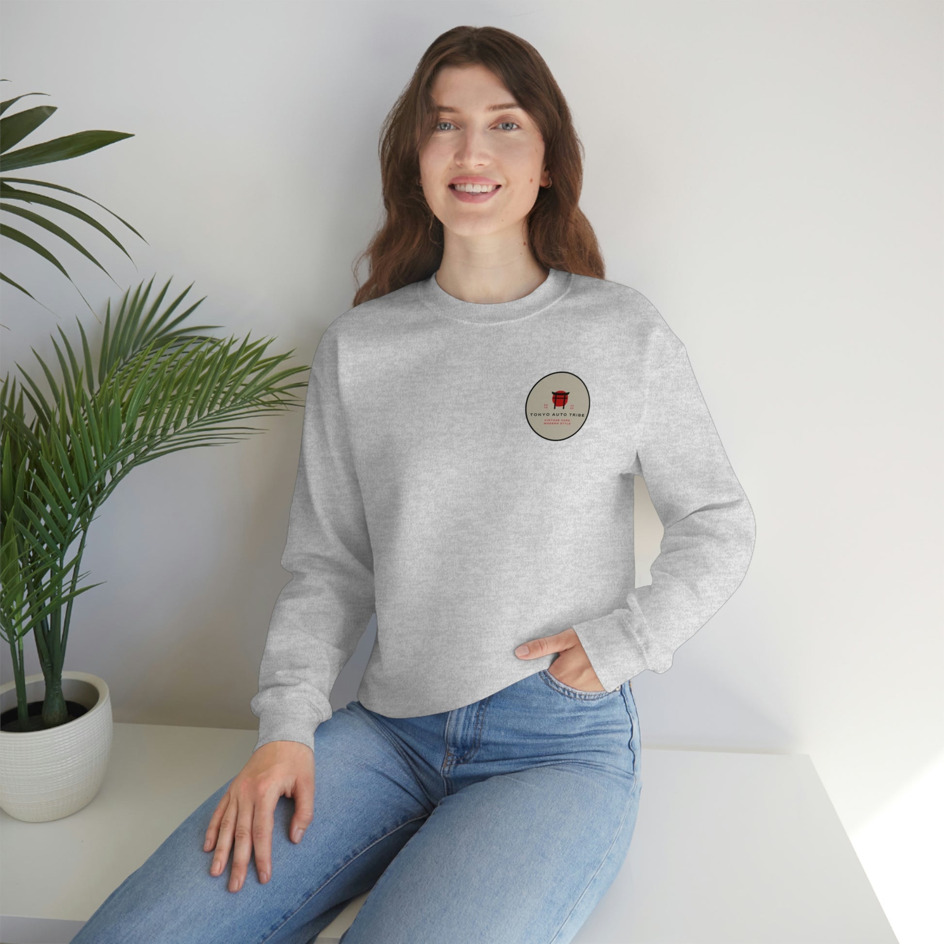 A woman wearing The Family Wagon Sweater, a light gray sweatshirt featuring a small circular logo with Japanese car culture-inspired design, styled casually with blue jeans.
