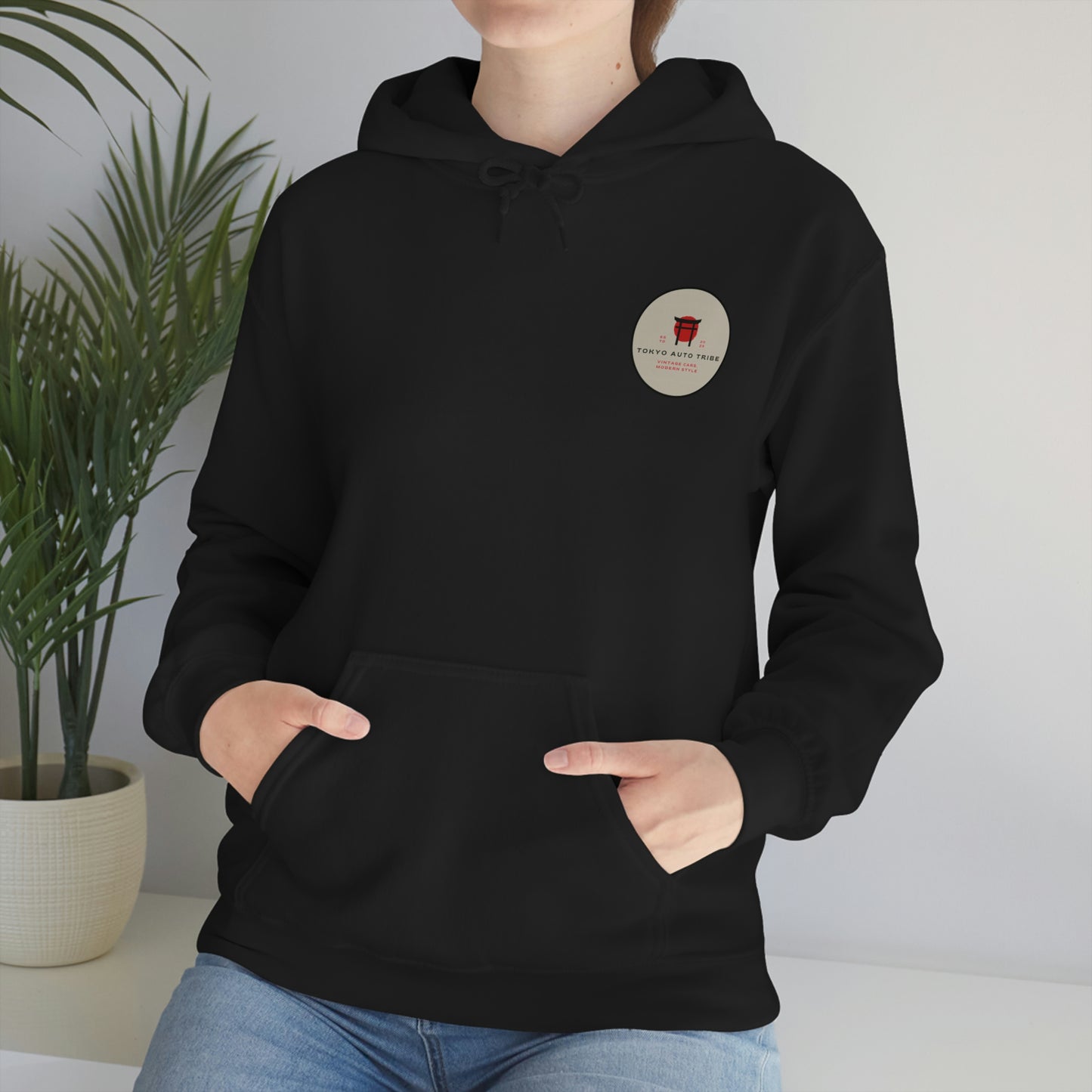 Person wearing a black Godzilla's Fury Hoodie with a small circular graphic on the chest, standing indoors near a green plant with hands in front pocket.